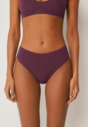 Frau trägt einen nahtlosen Bikini in Pflaumenfarbe, der den Oberkörper, die Arme und die oberen Beine vor neutralem Hintergrund zeigt.