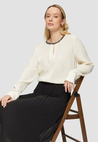 Weiße Bluse mit perlenbesetztem schwarzen Ausschnitt und Schlüssellochdetail, kombiniert mit einem schwarzen plissierten Rock. Model sitzt auf einem Holzstuhl.