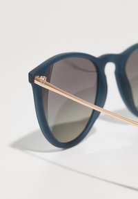 Ochelari de soare mat bluți cu o ramă rotundă, lentile întunecate cu un gradient și brațe subțiri din metal roz aurit cu un detaliu al logo-ului.