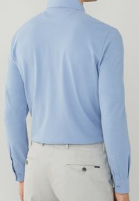 Uomo che indossa una camicia a maniche lunghe blu chiaro infilata in pantaloni beige chiaro, mostrato di spalle contro uno sfondo neutro.
