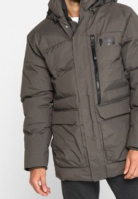 Giacca a vento in grigio scuro. Presenta una tasca sul petto con zip, due tasche frontali, un cappuccio e un design a pannelli cuciti. Materiale liscio e resistente all'acqua.