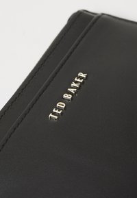 Crna kožna novčanik sa zlatnim reljefnim logom "TED BAKER", glatke teksture i kontrastnim šavovima duž ivica.
