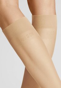 Beige transparente Oberschenkelstrümpfe mit einem strukturierten Band am oberen Ende, auf dem der Markenname "HUDSON" eingeprägt ist. Glatte Stoffoberfläche.