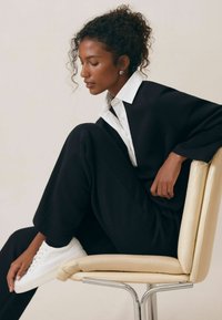 Čierna oversized mikina a zodpovedajúce nohavice, biela košeľa pod ňou, sediaca na chrómovo koženej stoličke, s bielymi teniskami.