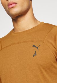 Brązowy t-shirt z okrągłym dekoltem, wykonany z miękkiego materiału. Posiada czarne logo puma oraz akcentujący wzór na klatce piersiowej z widocznymi detalami szwów.
