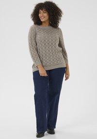 Lichtgrijze gebreide trui met ruitpatroon, ronde halslijn en driekwart mouwen, gecombineerd met donkerblauwe denim broek en zwarte schoenen.