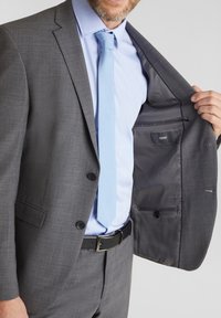 Giacca grigia con due bottoni, indossata aperta. Cravatta azzurro chiaro, camicia azzurro chiaro sotto. L'interno presenta una tasca e un tessuto liscio.