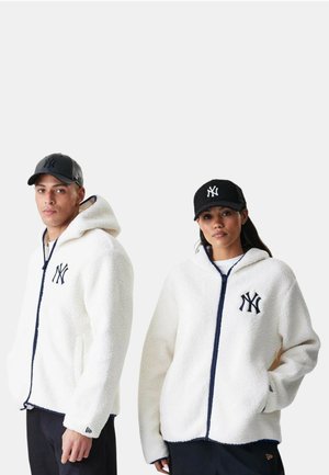 Mies ja nainen pukeutuneina valkoisiin fleecevetoketjuhuppareihin ja mustiin NY-logolla varustettuihin baseball-lippiksiin, seisomassa yksivärisen taustan edessä.