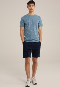 T-shirt bleu clair à manches courtes avec un motif d'étoiles blanches, associé à un short marine et des baskets légères, se tenant avec les mains dans les poches.