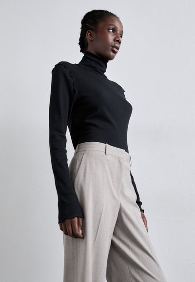 Sieviete, ģērbusies melnā pieguļošā turtleneck ar kraukšķīgiem malām un augsto jostasvietu beigu platā stila biksēs, stāv pretī vienkāršai siena.