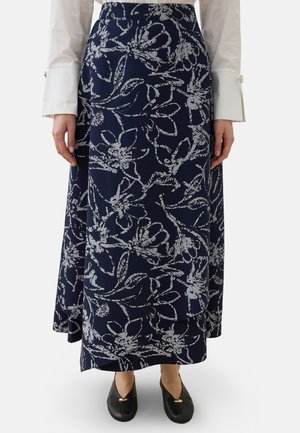 Femme portant une jupe longue bleu marine à motif floral, un chemisier blanc à larges poignets et des chaussures plates noires avec des détails dorés.