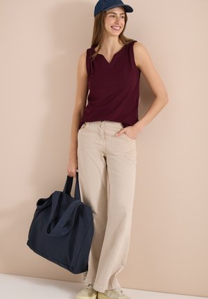 Jeune femme souriante, portant une casquette bleue, un haut bordeaux sans manches, un pantalon beige à jambes larges, tenant un grand sac cabas bleu marine.
