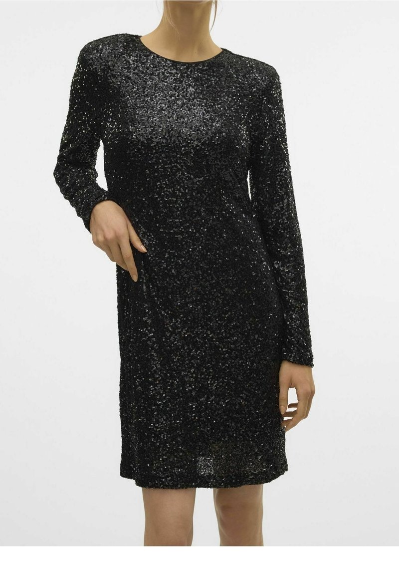 Femme portant une robe noire à manches longues, ornée de sequins, longueur genou, sur un fond blanc uni.