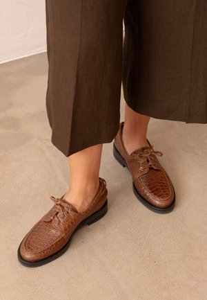 Zapatos marrones de cuero con textura y cordones, usados con pantalones cortos marrón oscuro sobre un suelo de color claro.