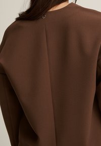 Braune Bluse mit glatter, matter Textur, rückseitigem Knopfverschluss und einem dezenten Nahtdetail, das durch die Mitte verläuft.