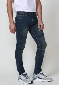 Vaqueros oscuros ajustados de color azul con secciones desgastadas y detalles acanalados en las rodillas. Combinados con zapatillas deportivas grises con acentos en color marino.