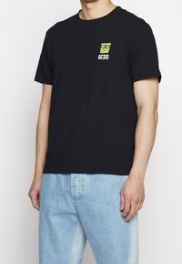 Černé bavlněné tričko s malým vyšitým designem s karikaturou obličeje a logem "GCDS" na hrudi. Standardní kulatý výstřih.