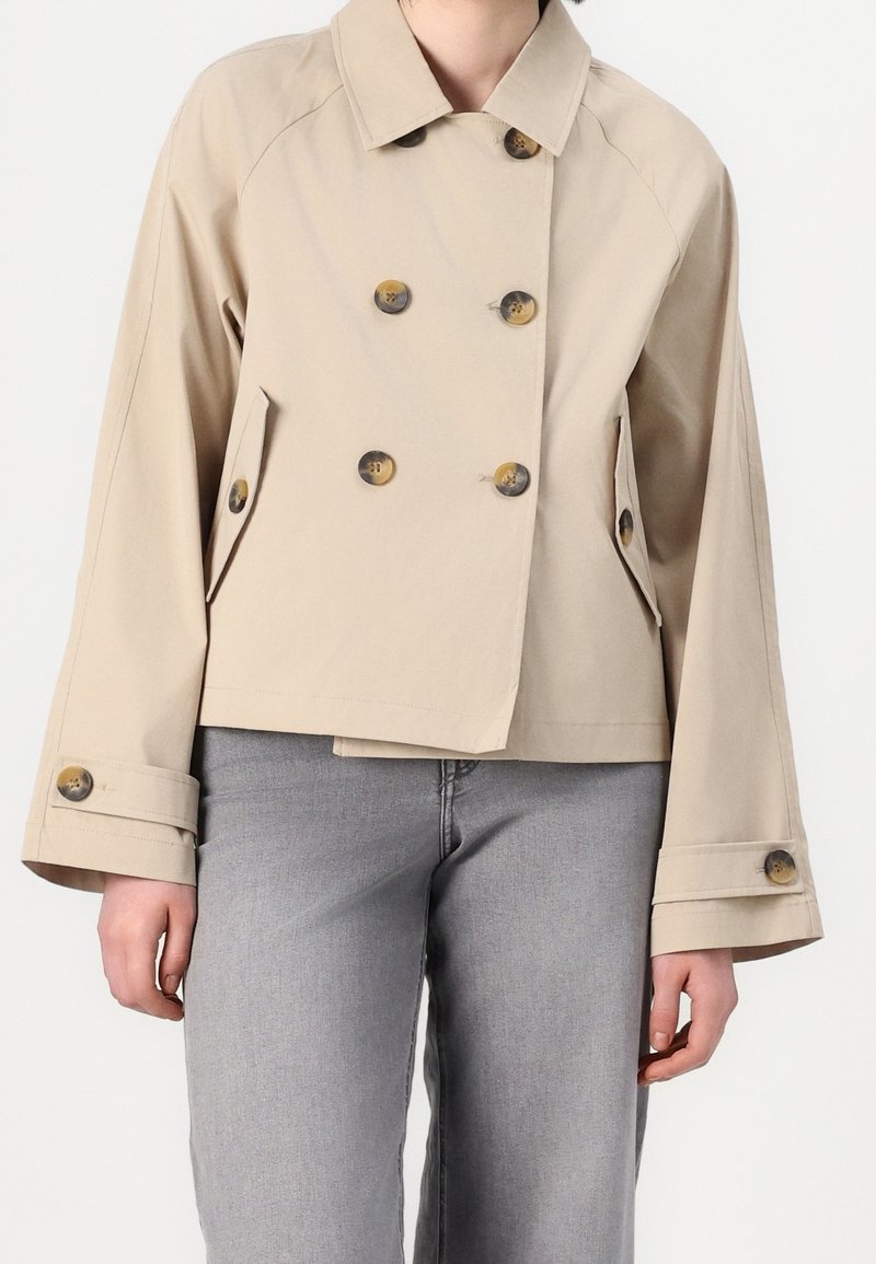 Veste courte beige à double boutonnage avec de grands boutons et des poches à rabat, portée avec un jean gris clair.