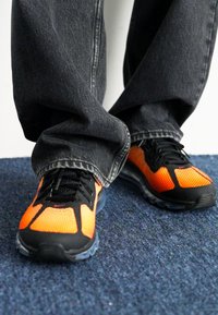 Zapatillas deportivas con parte superior de malla naranja, acentos negros y cordones visibles, combinadas con jeans grises holgados sobre una superficie de moqueta azul.