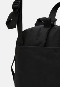 Sac à dos en tissu noir avec une surface texturée, des bretelles réglables et une poche latérale. Présente des éléments de design simples et fonctionnels.