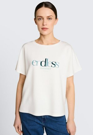 Kobieta z ciemnymi włosami związanymi z tyłu, ubrana w luźny biały T-shirt z napisem "endless summer" i niebieskie dżinsy, stoi na jasnym tle.