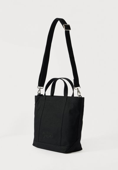 Čierna plátená taška na rameno s krátkymi rukoväťami a nastaviteľným dlhým popruhom, s jemnou výšivkou "Maison Kitsuné" na prednej strane.