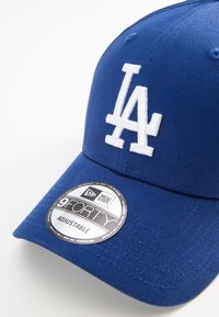 Căciulă de baseball albastră cu logo alb "LA", curea ajustabilă și țesătură texturată. Are un autocolant New Era 9FORTY pe bor.