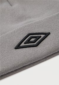 Skaidrs adīts audums ar melnu izšūtu dubultā dimanta logo uz salocīta malas, fona ir balts.