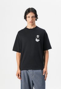 Black cotton t-shirt with short sleeves, featuring a small white graphic print on the chest pocket area. Relaxed fit design.