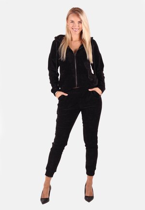 Frau lächelt, steht mit den Händen in den Taschen, trägt eine schwarze Kapuzenjacke mit Reißverschluss, passende Jogginghosen und schwarze High Heels.