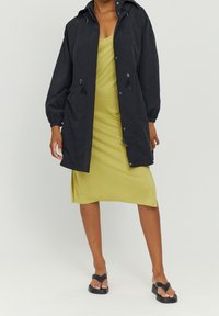 Czarna wodoodporna kurtka z troczkami, długimi rękawami i prostym krojem, noszona na oliwkowej sukience o długości do kolan oraz czarnych sandałach.