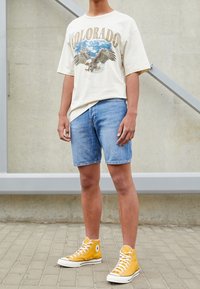 Camiseta gráfica beige con el texto "COLORADO" y estampado de águila; pantalones cortos de mezclilla azul; zapatillas altas amarillas con suelas blancas.