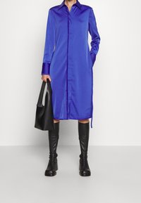 Modrá saténová košilová šaty s dlouhými rukávy, zapínáním na knoflíky vpředu a bočními kapsami, doplněné černými botami po kolena a černou kabelkou.