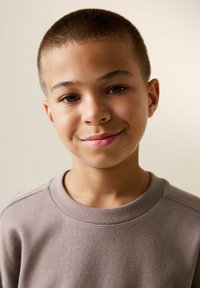 Sivý sveter s vaflovým vzorom a okrúhlym výstrihom, ktorý má na sebe chlapec s krátkymi vlasmi, usmievajúci sa na neutrálnom pozadí.