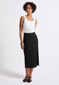 Jupe midi noir côtelée avec une fente sur le côté, associée à un top sans manches blanc. La tenue est agrémentée de bracelets ronds noir et blanc et de chaussures en toile noires.