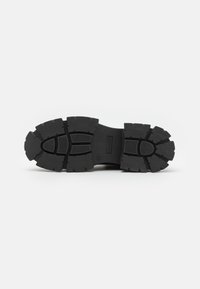 Suela de goma negra con surcos profundos y patrones texturizados. Forma plana y robusta diseñada para ofrecer tracción y durabilidad.