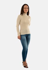 Hellbeige, gerippter Rollkragenpullover mit langen Ärmeln, kombiniert mit enganliegenden blauen Jeans und schwarzen, hohen Sandalen.