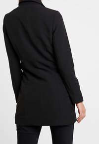 Czarny, dopasowany płaszcz o wyraźnej sylwetce, z długimi rękawami i klapami z wcięciem, charakteryzujący się gładką fakturą i ozdobnym szwem z tyłu.