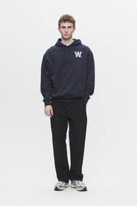 Giovane uomo con capelli ricci che indossa un hoodie blu navy con logo bianco, pantaloni neri e sneakers bianche, in piedi contro uno sfondo bianco.