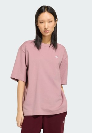 Jeune femme aux cheveux lisses et foncés porte un t-shirt oversize mauve clair et un pantalon bordeaux foncé, debout devant un fond uni.
