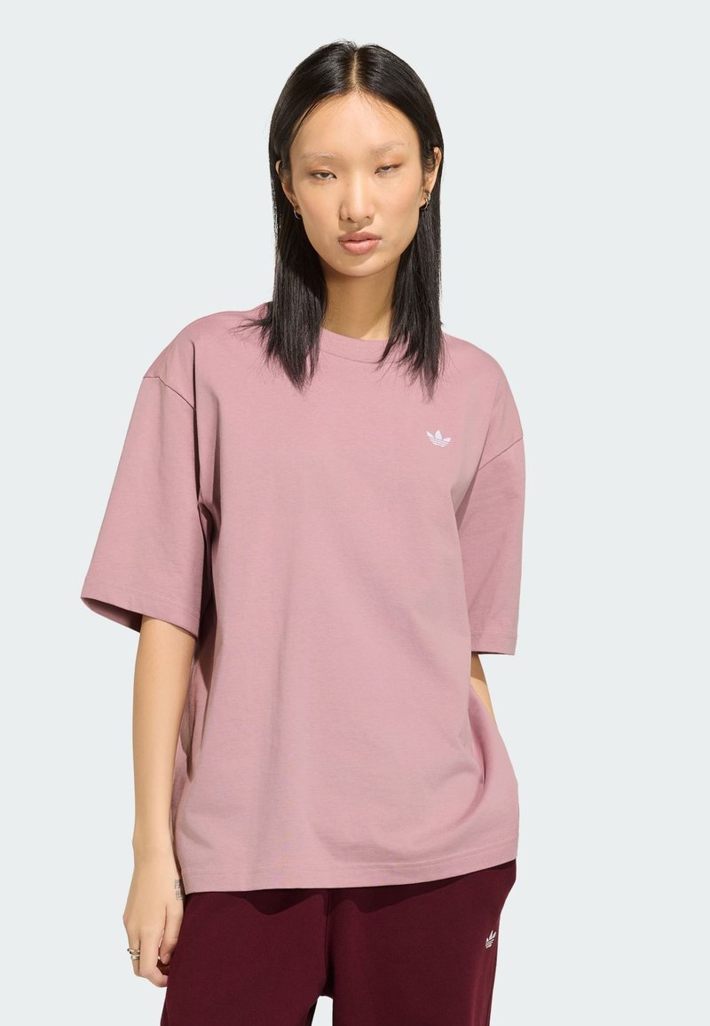 Jeune femme aux cheveux lisses et foncés porte un t-shirt oversize mauve clair et un pantalon bordeaux foncé, debout devant un fond uni.