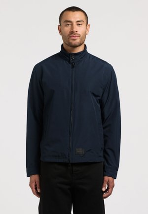 Hombre con cabello corto que lleva una chaqueta azul marino con cremallera, cuello con botones y pantalones negros, de pie frente a un fondo claro y liso.