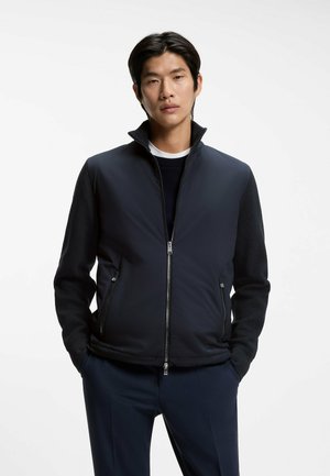 Ung mand står med front mod kameraet med hænderne i lommerne, iført mørk marineblå lynlåstrøje over sort langærmet trøje og matchende bukser.