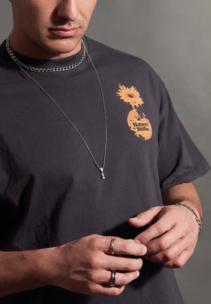 Black t-shirt featuring an orange pineapple graphic with flowers and text. Silver chain necklace, multiple rings, and textured bracelet details.