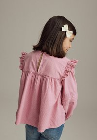 Lyserød bomuldsbluse med flæser på ærmerne, nøglehul bagpå, tekstureret stof og broderede detaljer på ærmerne, parret med blå denimjeans.