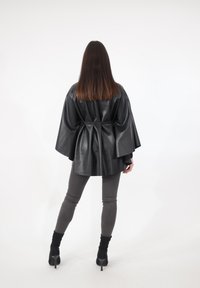 Czarna peleryna kurtka z ekoskóry o przemyślanej konstrukcji, z taliowanym pasem i luźnymi rękawami. Noszona z szarymi legginsami i czarnymi szpilkami.