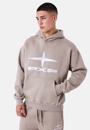 Joven con cabello corto vistiendo una sudadera con capucha beige y pantalones deportivos a juego con un logo de estrella blanca y el texto "PXP", de pie con las manos en los bolsillos.