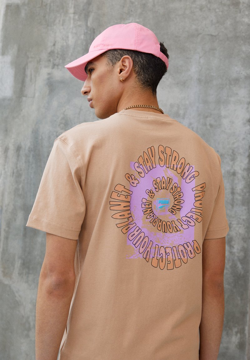 T-shirt en coton beige avec un texte graphique arrondi en violet et orange au dos, assorti à une casquette rose clair. Accent de chaîne en or.