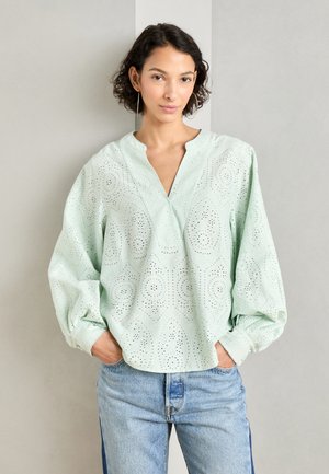 Blusa verde menta chiaro con dettagli a occhio di pernice, scollatura a V e maniche ampie. Abbinata a jeans blu chiaro.