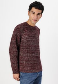 Bordeaux strikkegenser med teksturert mønster, rund hals og lange ermer, kombinert med blå jeans.